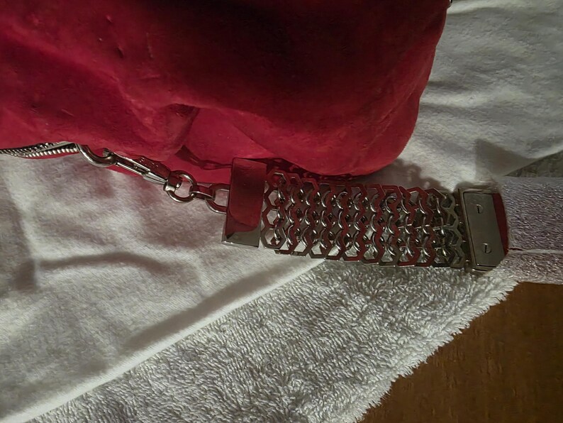 Puede incluir: Un bolso de mano rojo brillante con una superficie texturizada y una correa de cadena de metal plateado. La correa presenta un patr&oacute;n de eslabones geom&eacute;tricos repetitivos y se conecta al bolso mediante un cierre de metal plateado.