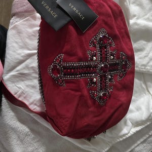 Puede incluir: Un bolso de mano de ante rojo intenso con un gran adorno de cruz decorado con metal oscuro, peque&ntilde;os cristales negros y piedras rojas facetadas m&aacute;s grandes. Dos etiquetas negras de la marca Versace est&aacute;n unidas arriba y se ve una correa de cadena.