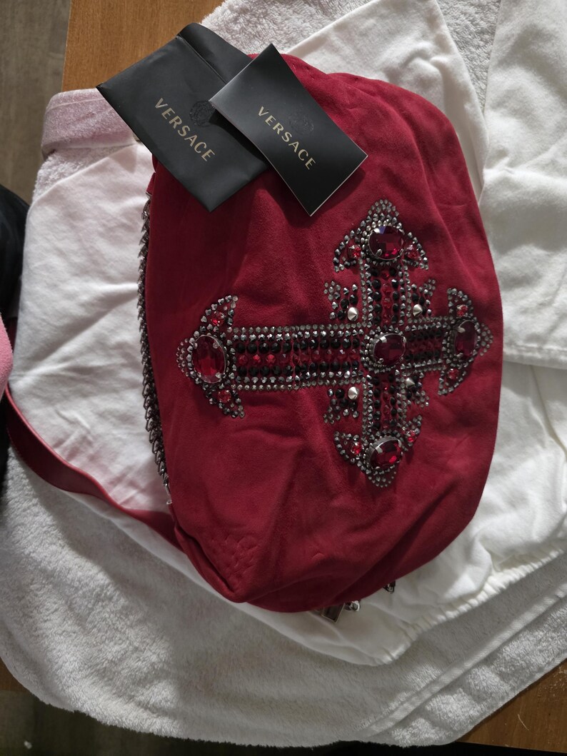 Puede incluir: Un bolso de mano de ante rojo intenso con un gran adorno de cruz decorado con metal oscuro, peque&ntilde;os cristales negros y piedras rojas facetadas m&aacute;s grandes. Dos etiquetas negras de la marca Versace est&aacute;n unidas arriba y se ve una correa de cadena.