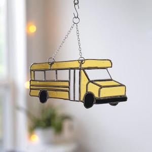May include: A yellow stained glass suncatcher shaped like a school bus, featuring black glass wheels and dark metal framing. The bus is suspended by a silver-toned metal chain and an S-hook, allowing it to hang vertically for display in a window.