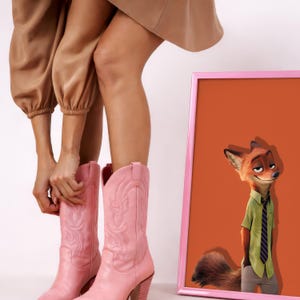 May include: A pair of light pink leather cowboy boots featuring intricate tonal stitching on the shaft and a stacked heel, positioned next to a framed rectangular print displaying a cartoon fox character wearing a green shirt and a dark tie.