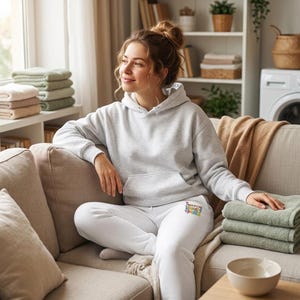 May include: A light gray hooded sweatshirt with long sleeves and a front kangaroo pocket, paired with white sweatpants featuring a small, colorful graphic print on the left thigh that reads "I love laundry day".