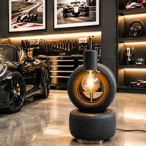 May include: A floor lamp constructed from two stacked black rubber racing tyres. The upper tyre features a central vertical metal rod holding a single glowing Edison-style light bulb. The base of the lamp rests on a small wooden block on the floor.