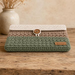 May include: A rectangular crochet clutch bag featuring three horizontal color-blocked sections in cream, tan, and sage green. The flap is secured with a single brown wooden button and a loop closure. A small rectangular leather tag reads handmade.