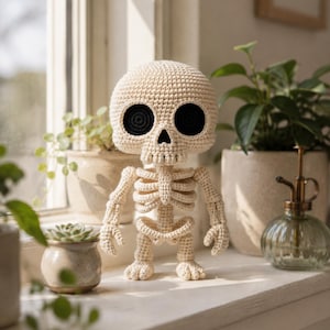 Op de afbeelding: Een crèmekleurig gehaakt skeletfiguur met een grote, ronde schedel, diepzwarte ronde oogkassen, een driehoekige neusopening en een ribbenkaststructuur. Het figuur heeft beweegbare armen en benen met eenvoudige, ronde handen en voeten.