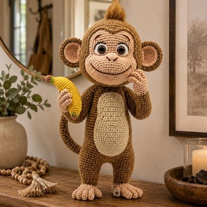 Puede incluir: Un juguete de mono tejido a ganchillo con hilo marrón, que presenta una barriga de color beige claro, orejas grandes y un mechón de pelo marrón en la cabeza. El mono tiene ojos grandes, una expresión sonriente y sostiene un plátano amarillo.