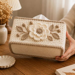 May include: A rectangular tissue box cover made of cream-colored crochet yarn. The front panel features a central three-dimensional crocheted flower in cream and beige, flanked by crocheted leaves and small round buds in a light brown yarn.