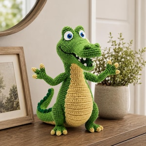 Op de afbeelding: Een gehaakt alligator-speeltje in groen en geel met een textuurrijk oppervlak, voorzien van grote blauw-witte ogen, een open mond met zichtbare witte tanden, opgeheven armen en een lange staart. Het wezen is gemaakt met gedetailleerd garen.