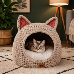 Pode incluir: Uma cama de gato em crochê bege com formato de cabeça de gato e orelhas pontudas com detalhes em rosa. A cama tem uma abertura circular com uma almofada de pelúcia branca onde descansa um gato castanho e branco de olhos azuis. Uma etiqueta castanha diz Handmade with love.