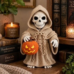 Op de afbeelding: Een gehaakt skeletfiguur in een beige gewaad met capuchon, die een kleine oranje pompoen vasthoudt. Het skelet heeft een witte schedel met zwarte oogkassen en een gestikte mond, en het gewaad heeft een koord rond de taille.