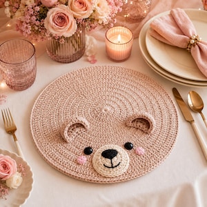 May include: A round, light pink crocheted placemat featuring a bear face design. The placemat has two small, rounded ears, two black circular eyes, a cream-colored muzzle with a black embroidered nose and mouth, and two small pink cheeks.