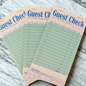 May include: Three rectangular paper guest check pads with a light tan header and a pale green body. The header features the blue text Guest Check, with fields for date, table, guests, and server. The green section contains lined rows for orders.