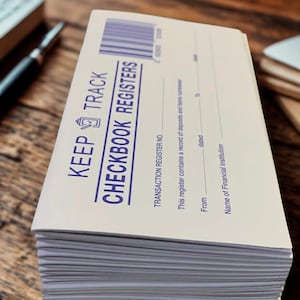 May include: A stack of off-white checkbook registers with blue text and a barcode. The cover reads KEEP TRACK CHECKBOOK REGISTERS and includes fields for transaction register number, date range, and the name of the financial institution.