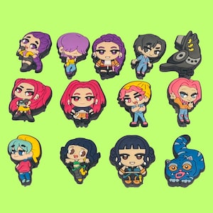 May include: A collection of fourteen colorful rubber shoe charms featuring various cartoon-style characters, including figures with purple, pink, and black hair, a dark grey mechanical creature, and a blue striped cat with large eyes.