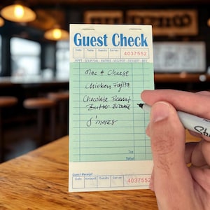 以下が含まれることがあります： 上部に「Guest Check」と記された白いセクションと、薄緑色の罫線が入った本体を持つ縦型のゲストチェックパッド。手書きのテキストには、黒インクでMac & Cheese、Chicken Fajita、Chocolate Peanut Butter Brownie、S'moresと記載されています。