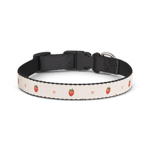 Può includere: Un collare per animali domestici con un cinturino color crema caratterizzato da un motivo ripetuto di piccole fragole rosse e minuscoli cuori rosa. Il collare include una fibbia a scatto in plastica nera, un cinturino di regolazione nero e un anello a D in metallo argentato.