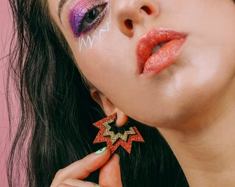 May include: A woman with colorful eye makeup and a red glitter star-shaped earring. The earring is made of two layers of red glitter with a gold glitter layer in between.