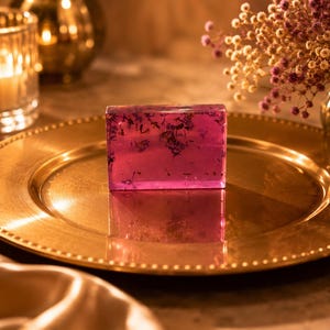 May include: A rectangular bar of translucent magenta soap embedded with small dried purple flower petals. The soap has a smooth, glossy finish and is positioned upright on a polished gold-toned plate with a beaded rim, reflecting the light.