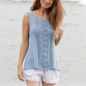 May include: A sleeveless, light blue knitted top with a detailed central panel and a V-neck. The top is paired with white shorts with a lace trim. The image is set against a blurred beach background. The logo for Drops Design is visible.