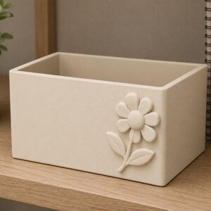 Può includere: Un contenitore rettangolare color beige con una finitura opaca e liscia. Il lato anteriore presenta un rilievo tridimensionale di un singolo fiore con un disco centrale circolare, otto petali arrotondati e due foglie su un gambo corto.