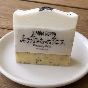 May include: A rectangular bar of soap with a white top layer and a pale yellow bottom layer, both containing small black poppy seeds. A paper label wraps around the center, printed with the text Lemon Poppy, Blackberry Betty, and net weight 4 oz.