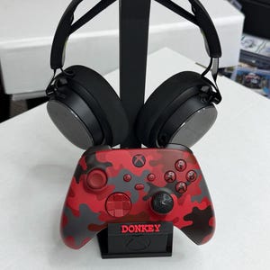 May include: A black gaming headset resting on a vertical stand above a red, gray, and black camouflage patterned wireless game controller. The controller features the word DONKEY in red lettering on a small black base with an Xbox logo emblem.
