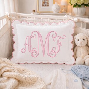 May include: A rectangular white pillow featuring a scalloped edge trimmed in pink thread. The centre of the pillow displays a large, decorative embroidered monogram in pink thread with the letters A, M, and F in a flowing, cursive script style.