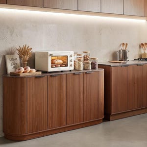 May include: A kitchen sideboard with a fluted wood finish and a dark countertop. On top sits a cream-colored toaster oven, a wooden tray with three donuts, a dried wheat arrangement, and a book titled LIGHT AND SALT LIFE, alongside glass jars.