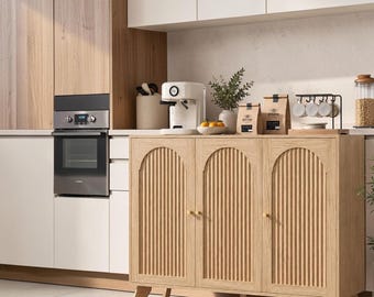 May include: A light oak sideboard cabinet featuring three doors with vertical fluted paneling and arched top details. The cabinet stands on four tapered legs and includes small, round, gold-toned knobs on each door for opening.
