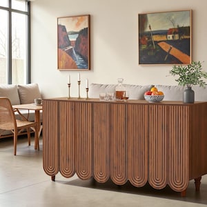 May include: A rectangular wooden sideboard featuring a dark brown finish and a scalloped base design with vertical grooved lines. The top surface holds three brass candlesticks, two clear glass tumblers, a glass decanter, and a bowl of fruit.