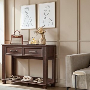 May include: A dark brown wooden console table with two drawers and a lower shelf. On top sits a beige and brown handbag, a glass diffuser, and a textured vase with dried flowers. Two framed line art prints hang above the table on a paneled wall.