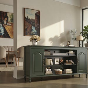 May include: A dark green wooden media console featuring two side cabinets with arched panel doors and ornate metal handles, plus an open central shelving unit with three tiers holding books, decorative items, and a small basket.