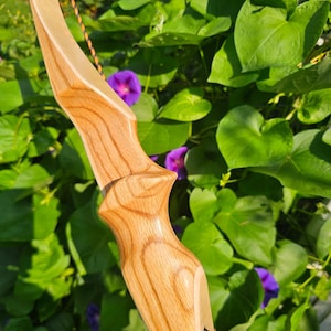 May include: A recurve bow featuring a light-colored wood riser with a prominent grain pattern and a smooth, polished finish. The bow includes a black and orange braided string attached to the limbs, with black text printed on the lower riser section.