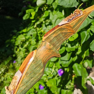 May include: A recurve bow riser crafted from laminated wood featuring a distinct green and brown grain pattern. The handle section includes two decorative inlays of textured, light-colored antler material positioned at the top and bottom.