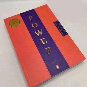 May include: A paperback book titled The 48 Laws of Power by Robert Greene. The cover features a bright orange background with a vertical purple stripe in the center. The word POWER is printed in large gold letters. A gold seal says National Bestseller.