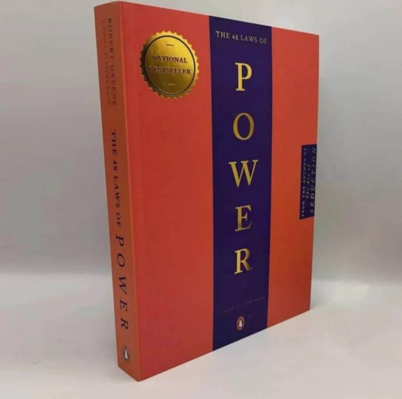 May include: A hardcover book titled The 48 Laws of Power by Robert Greene. The cover features a vibrant orange background with a central vertical purple stripe. Gold lettering displays the title, and a gold seal reads National Bestseller.