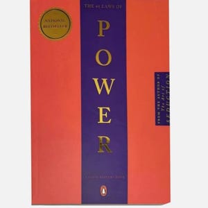 May include: A book titled The 48 Laws of Power with a bright orange cover and a central vertical purple stripe. The title is printed in gold lettering. A gold circular National Bestseller seal is in the top left corner, and a small penguin logo is at the bottom.