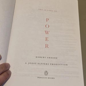 May include: The title page of the book The 48 Laws of Power by Robert Greene, a Joost Elffers production. The word POWER is printed vertically in red capital letters, with the Penguin Books logo and name at the bottom of the white page.