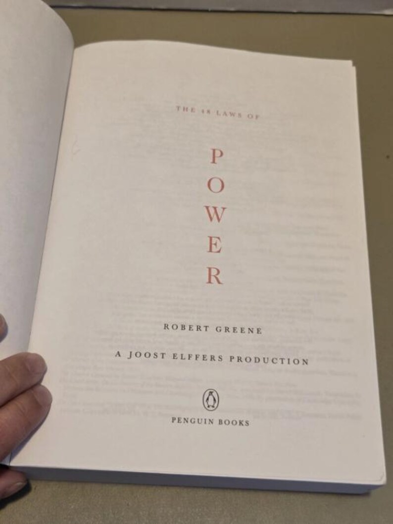 May include: The title page of the book The 48 Laws of Power by Robert Greene, a Joost Elffers production. The word POWER is printed vertically in red capital letters, with the Penguin Books logo and name at the bottom of the white page.