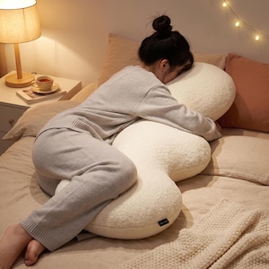 Puede incluir: Una almohada corporal grande de color blanco roto con una forma curva e irregular. La almohada presenta una superficie de tela suave similar al forro polar y una pequeña etiqueta de marca negra rectangular unida al borde lateral.