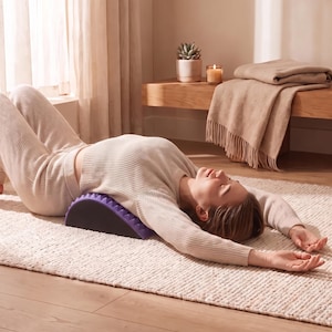 May include: A purple back stretcher device with a textured, spiked surface positioned under the upper back. The device has a semi-circular arch shape and a black base, designed to support the spine while lying flat on the floor.