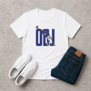 May include: A white short-sleeved t-shirt featuring a graphic of a football player in a blue uniform making a one-handed catch. The player is positioned over large blue letters that spell out OBJ. The shirt is paired with dark blue denim jeans.