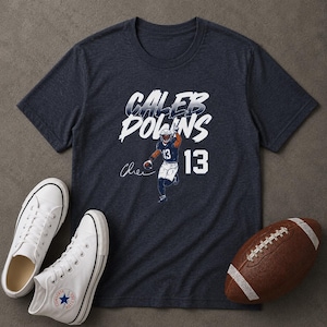 May include: A dark heather navy blue short-sleeve t-shirt featuring the white text CALEB DOWNS, a graphic of a football player in a blue uniform with the number 13, a signature, and the number 13 in large white font on the right side.