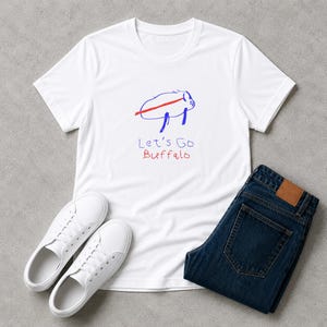May include: A white short-sleeved t-shirt featuring a hand-drawn blue buffalo outline with a red horizontal line across its body. Below the graphic, the text reads Let's Go Buffalo in blue and red handwritten lettering. The shirt is paired with dark blue denim jeans and white sneakers.