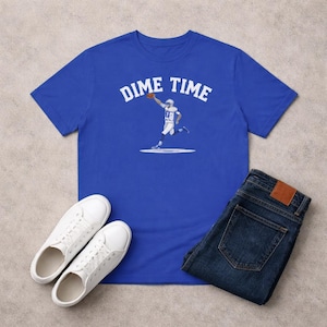 May include: A royal blue short-sleeve t-shirt featuring the white text DIME TIME arched above a graphic of a football player in a white uniform throwing a brown football. The shirt is paired with dark blue denim jeans and white sneakers.