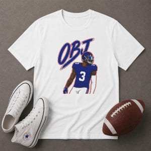 May include: A white short-sleeved t-shirt featuring a graphic of a football player wearing a blue jersey with the number 3 and a helmet, with the text OBJ in blue and red above the player. The shirt is displayed with white sneakers and a football.