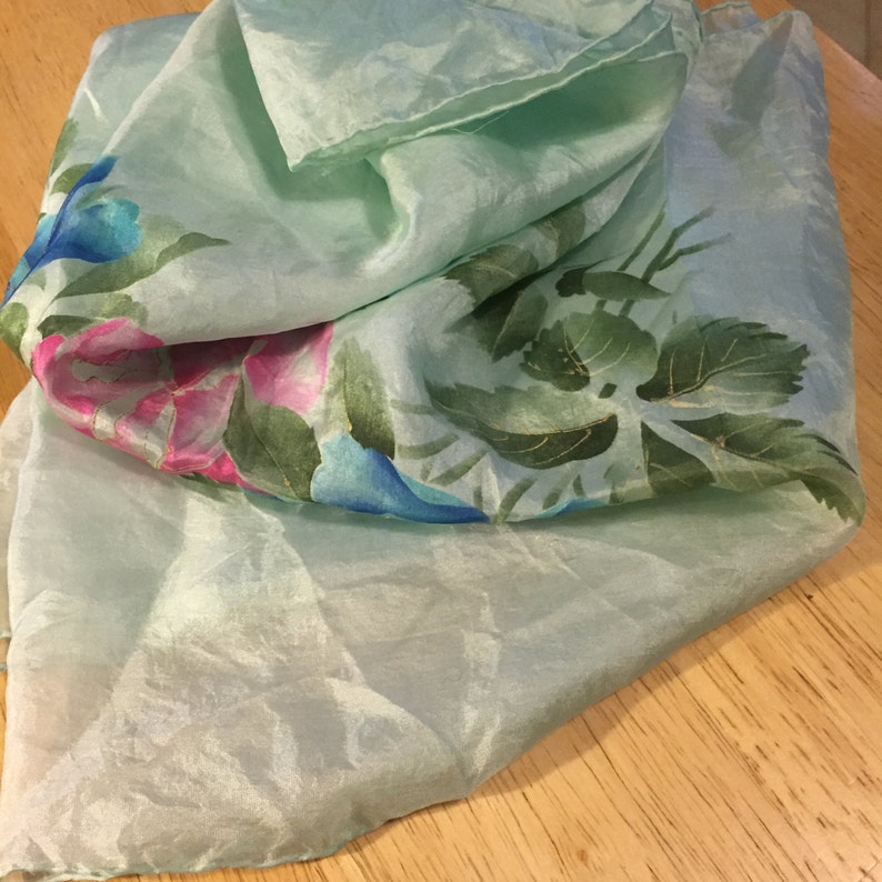May include: A light blue silk scarf with a floral print featuring pink, blue, and green flowers. The scarf is folded and draped on a wooden surface.