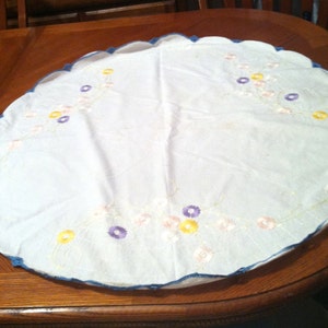 Puede incluir: Un mantel redondo blanco con un borde festoneado azul y dise&ntilde;os florales bordados en tonos amarillos, rosas y morados.