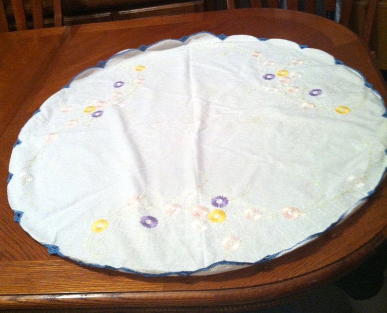 Puede incluir: Un mantel redondo blanco con un borde festoneado azul y dise&ntilde;os florales bordados en tonos amarillos, rosas y morados.