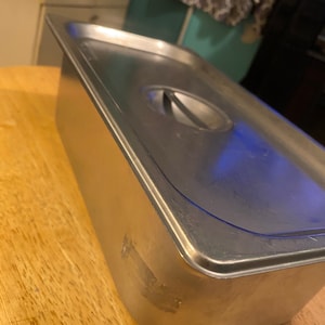May include: A stainless steel food pan with a lid. The pan has a slightly rounded edge and a small handle on the lid.